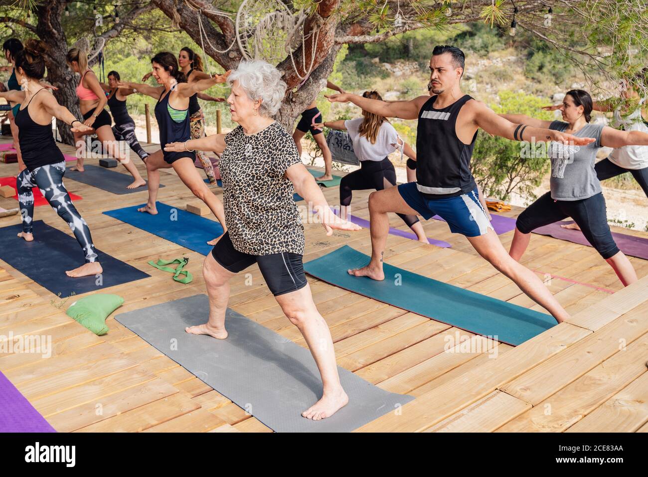 Gruppo di subacquei motivati persone in età diverse che allungano le gambe E caviglie mentre si esegue Virabhadrasana B posa durante lo yoga all'aperto classe Foto Stock