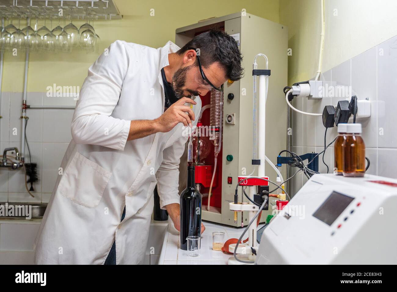 Scienziato maschile adulto in uniforme e occhiali che misura il contenuto di zucchero di vino mentre si usa un misuratore di vino trasparente in posizione di laboratorio vicino all'apparecchiatura elettronica Foto Stock