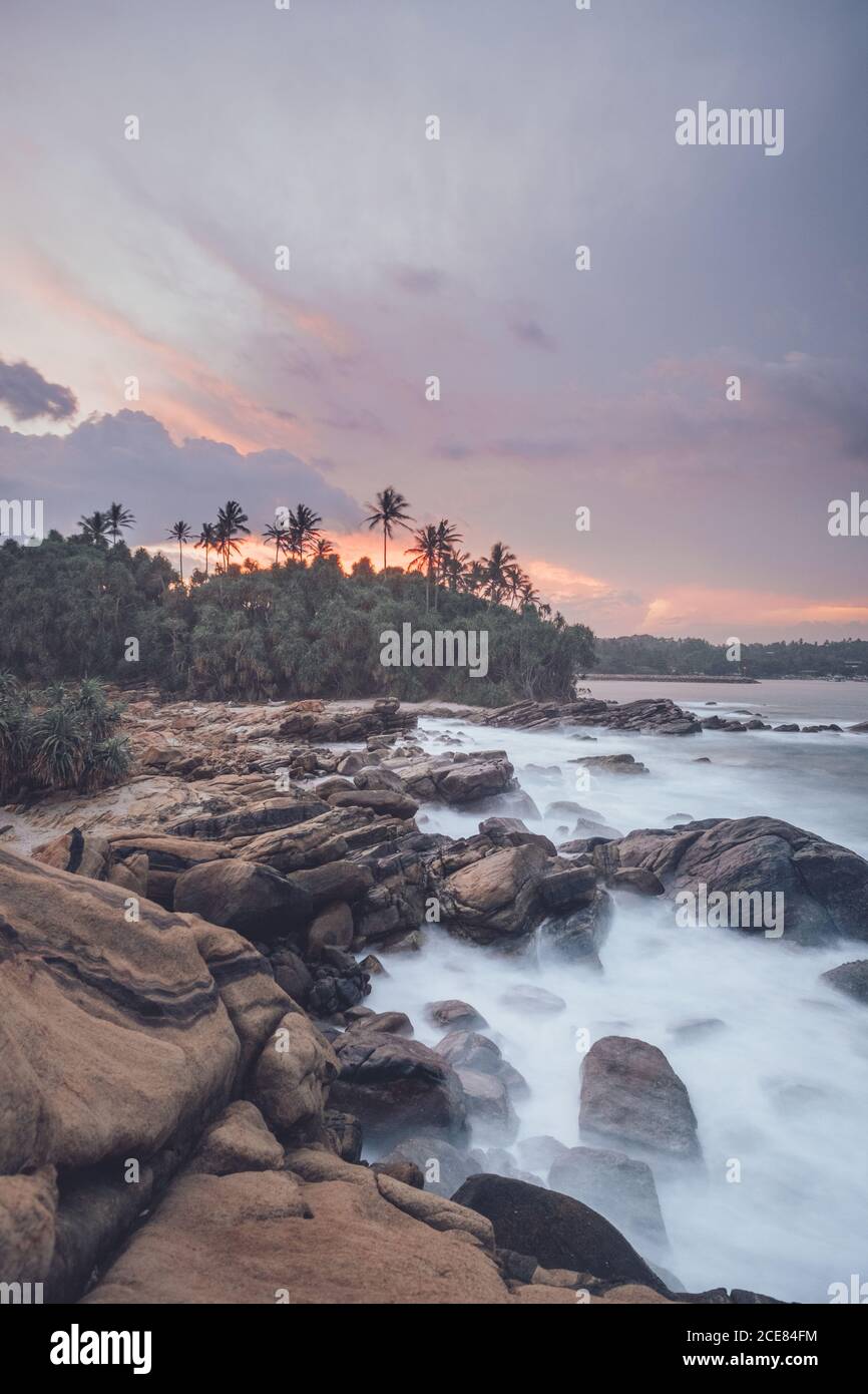 Incredibile scenario di costa rocciosa e palme verdi vicino mare sotto il pittoresco cielo del tramonto Foto Stock