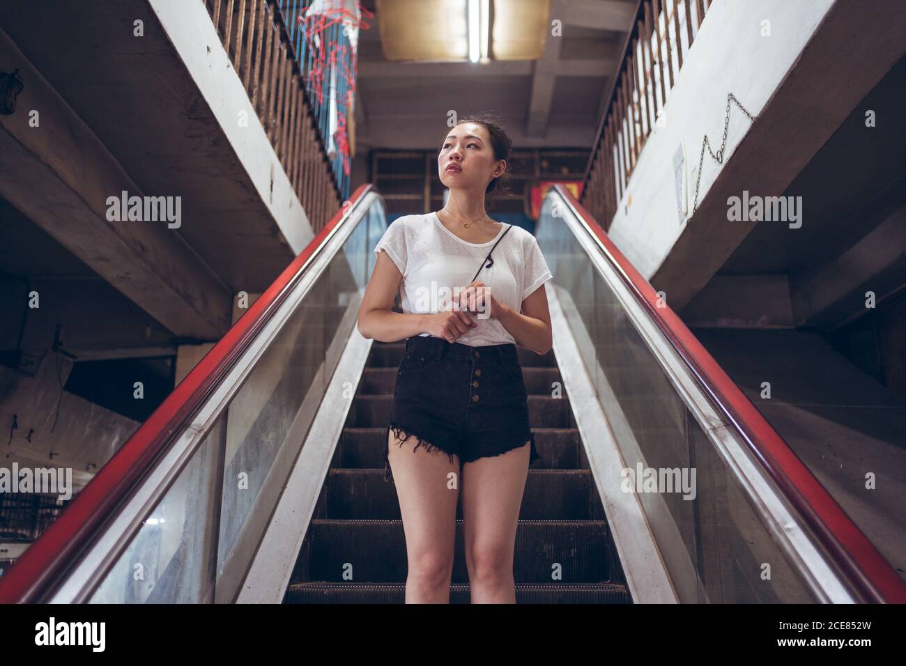Basso angolo di seria giovane donna asiatica in abbigliamento casual stare in piedi da soli sulla scala mobile scendendo in moderno edificio cittadino Foto Stock