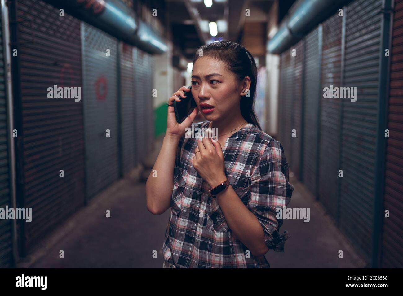 Infelice paura giovane donna asiatica in abbigliamento casual che hanno chiamata sul telefono cellulare mentre si è in piedi in un corridoio sotterraneo scuro grunge Foto Stock