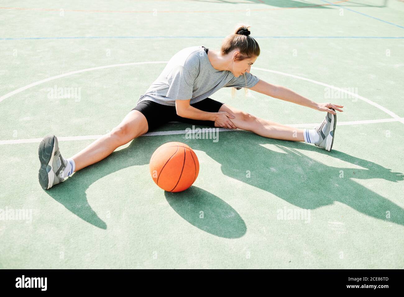 Vista ad alto angolo del corpo della giovane giocatrice di basket femminile seduto sul campo vicino alla palla e facendo esercizio di stretching prima gioco Foto Stock