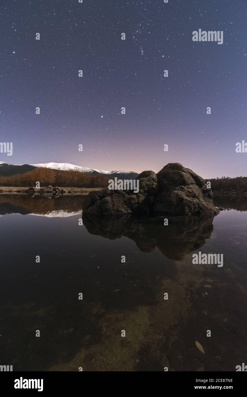 Incredibile scenario di lago e montagne innevate sotto il cielo scuro con stelle e una roccia in primo piano Foto Stock