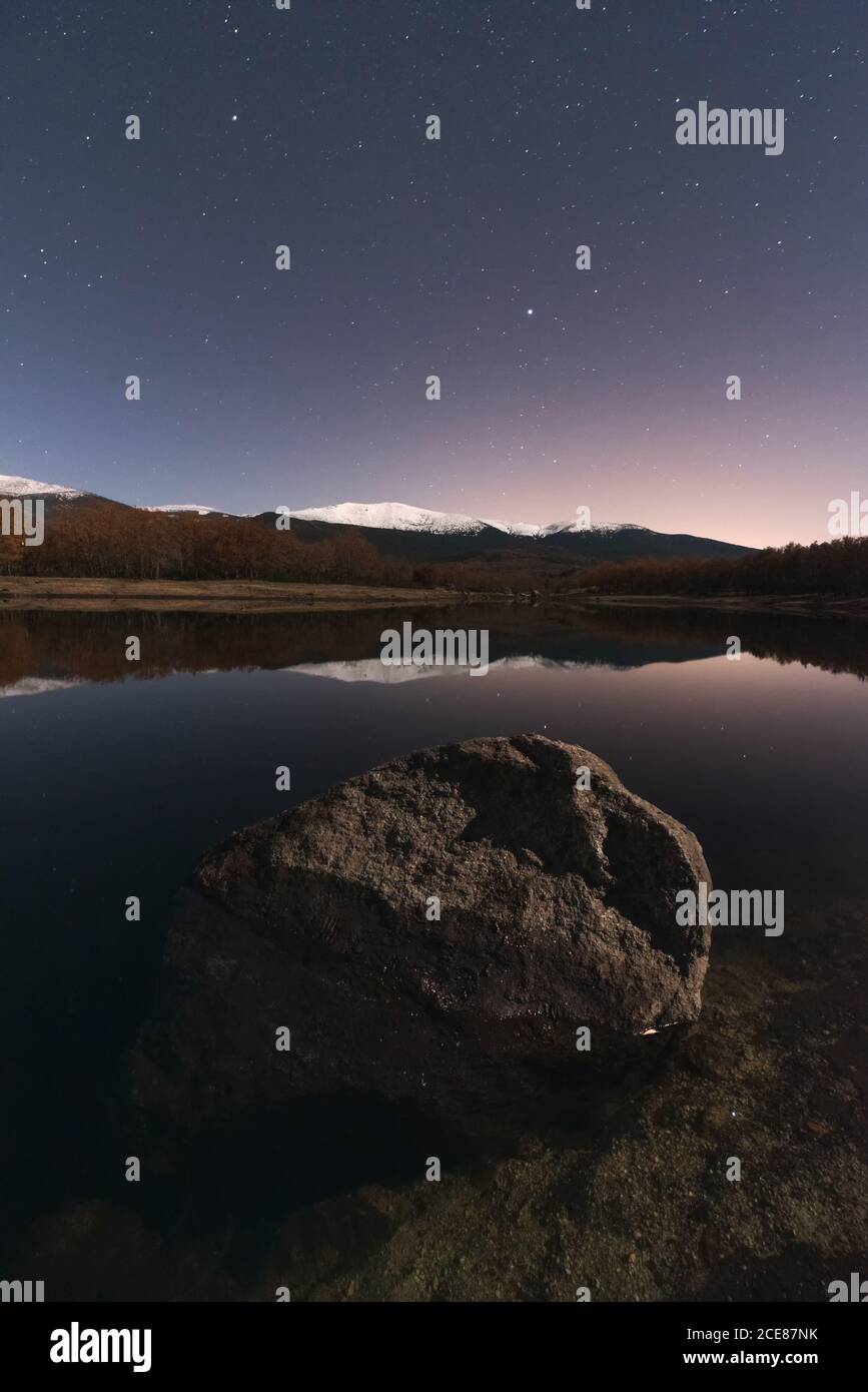 Incredibile scenario di lago e montagne innevate sotto il cielo scuro con stelle e una roccia in primo piano Foto Stock
