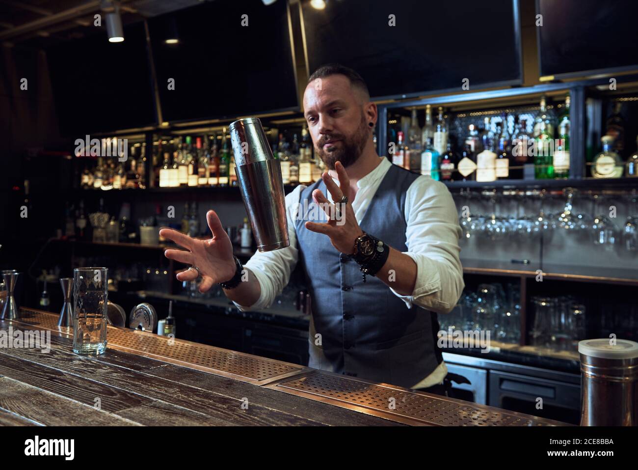 Buon barman professionista in elegante muta che mescola cocktail in shaker mentre si lavora al bar guardando lontano Foto Stock