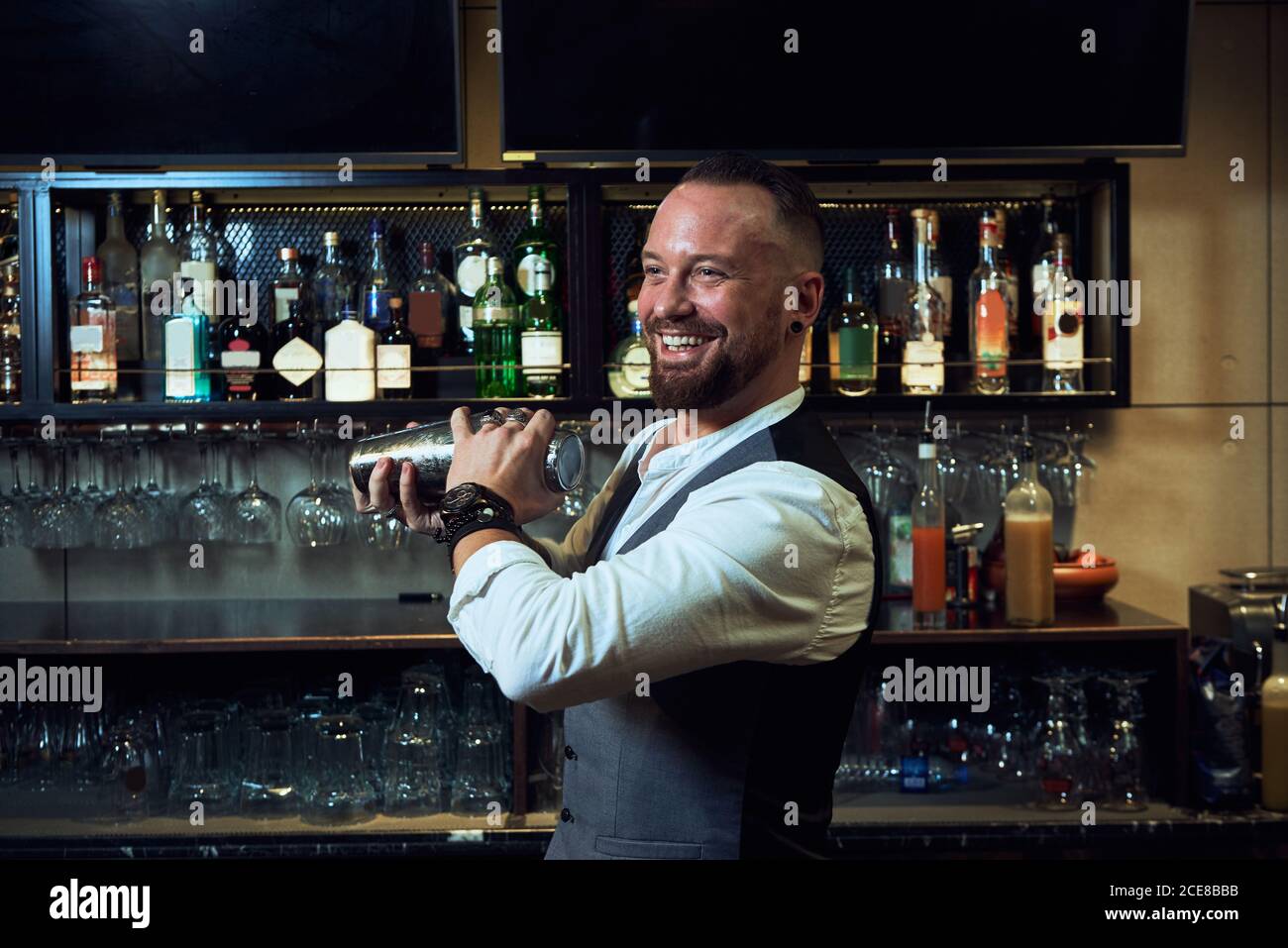Barman professionista allegro in elegante abito che mescola cocktail in shaker mentre si lavora al bar guardando lontano Foto Stock
