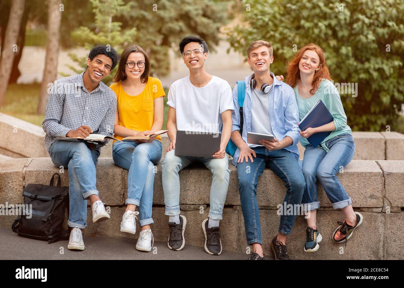 Felici e diversi studenti che posano seduti insieme all'esterno, sorridendo a Camera Foto Stock