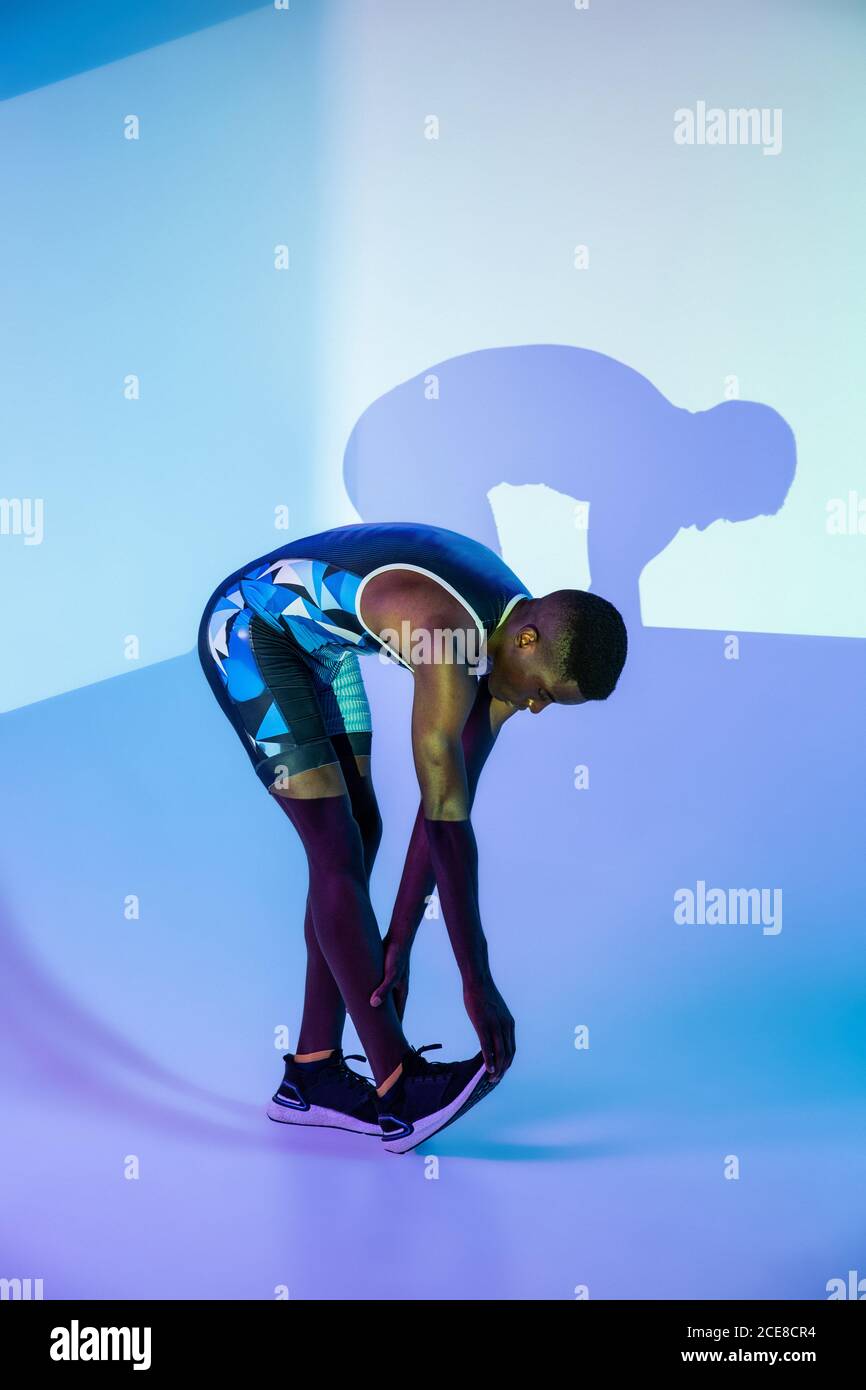 Vista laterale del corpo di un determinato professionista afroamericano maschio sprinter in tuta sportiva piegandosi in avanti e allungando le gambe prima inizia in studio con l'illuminazione al neon blu Foto Stock