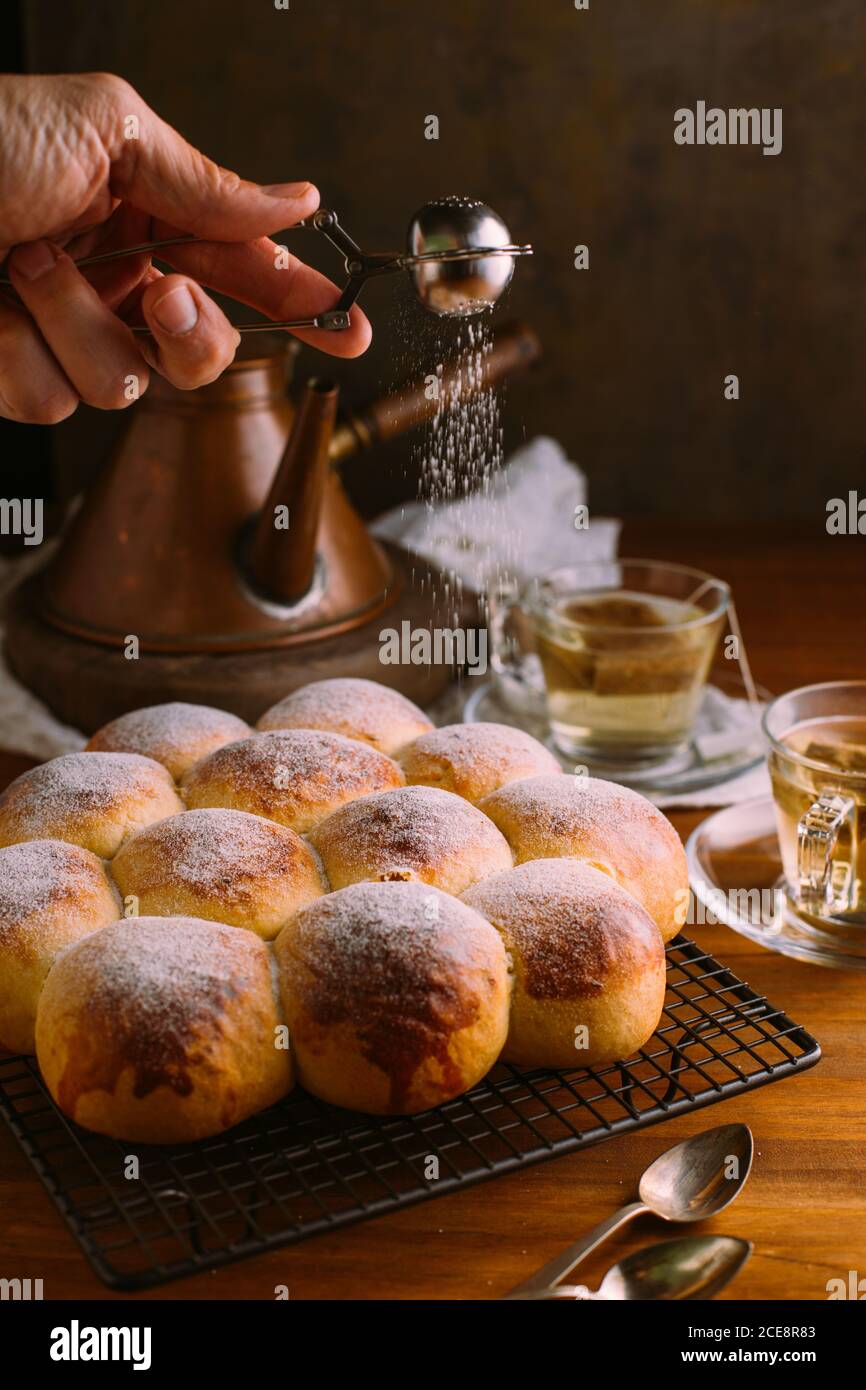 Crop anonimo chef setacciare zucchero a velo utilizzando un piccolo setaccio metallico mentre decorano i panini dorati gustosi vicino alle tazze con il tè verde borse sul tavolo Foto Stock