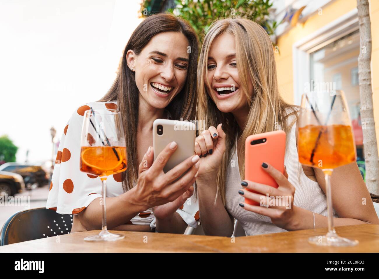 Immagine di due donne adulte sorridenti e che usano i telefoni cellulari mentre si bevono dei cocktail al tavolo in un caffè all'aperto Foto Stock