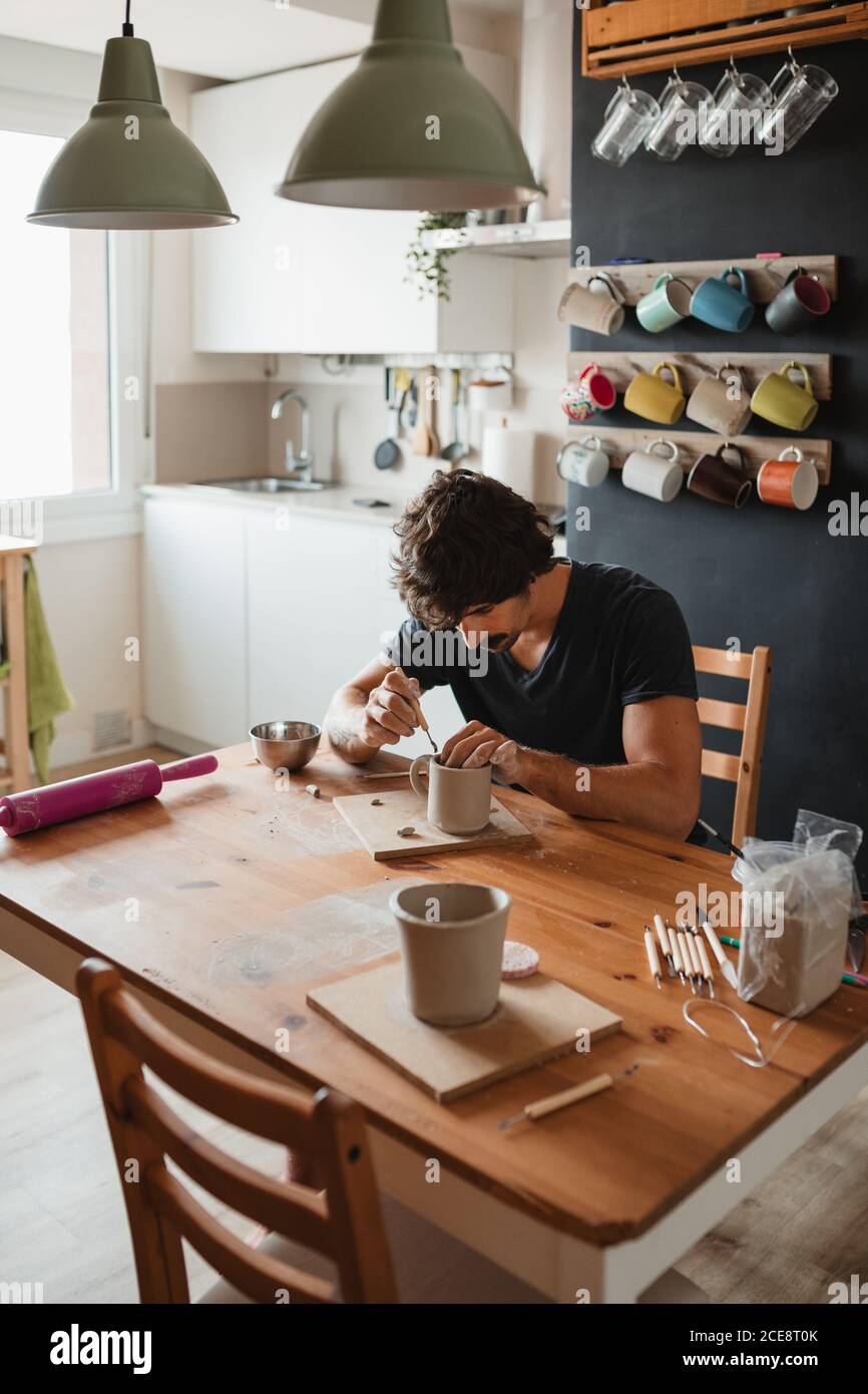 Artigiano seduto al tavolo di legno in officina e facendo tazza da creta Foto Stock