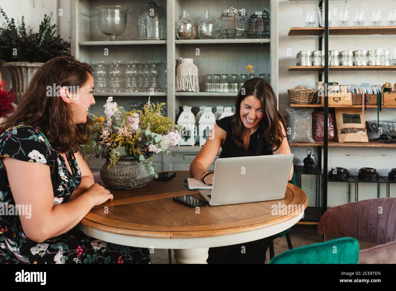 Vista laterale della donna proprietario di elegante salone di floristry discutere dettagli dell'ordine con il cliente seduto insieme al tavolo con il notebook in un atteggiamento felice Foto Stock