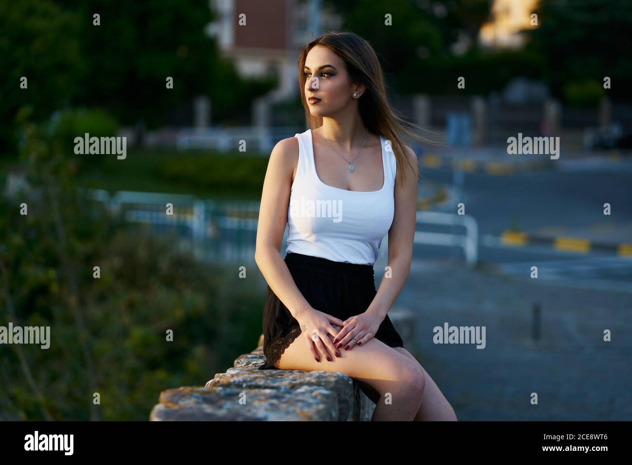 Donna pacifica in estate indossare seduta su recinzione di pietra su street e ammirare il tramonto incredibile Foto Stock