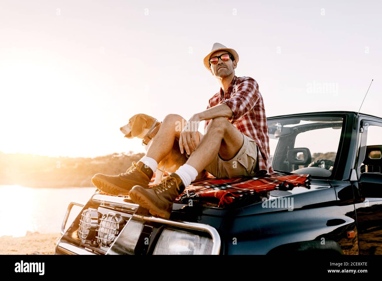 Sognante viaggiatore maschile in cappello e occhiali da sole seduti in auto vicino cane purebred con lingua fuori mentre si ammira il fiume e. montagne in giornata di sole Foto Stock