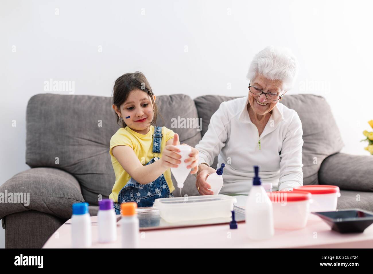 Sorridente erboso in occhiali aiutando nipote positiva con faccia sporca in abbigliamento casual preparazione di una miscela con coloranti in contenitore di plastica mentre si è seduti su un comodo divano nel soggiorno Foto Stock