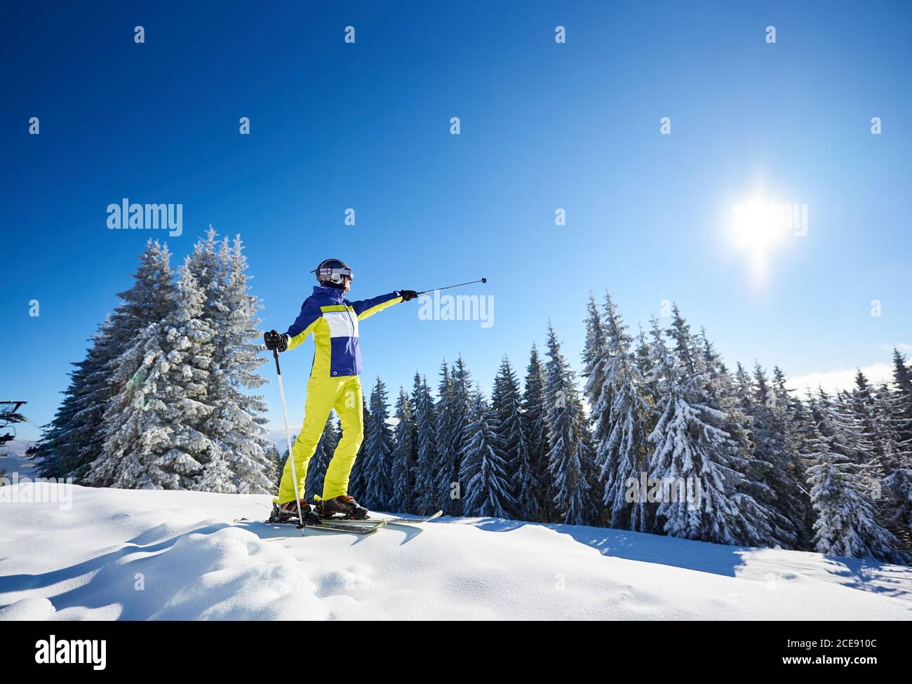 Foto a basso angolo di sciatore femminile prima di sciare da discesa. Donna sorridente in occhiali che mostra con pali da sci su sole luminoso in cielo blu. Alberi di abete innevati sullo sfondo. Stile di vita ricreativo. Foto Stock