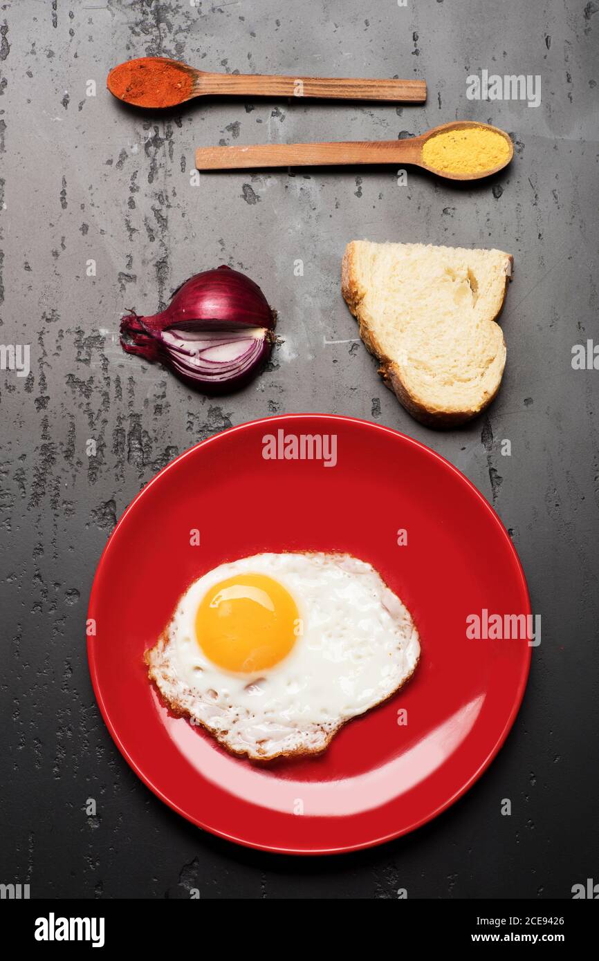 Omelette messa al centro del piatto. Colazione e concetto di arte alimentare. Condire con uova fritte vicino a cucchiai con spezie, metà fetta di pane e metà cipolla. Cibo posto su piatto rosso su fondo nero Foto Stock