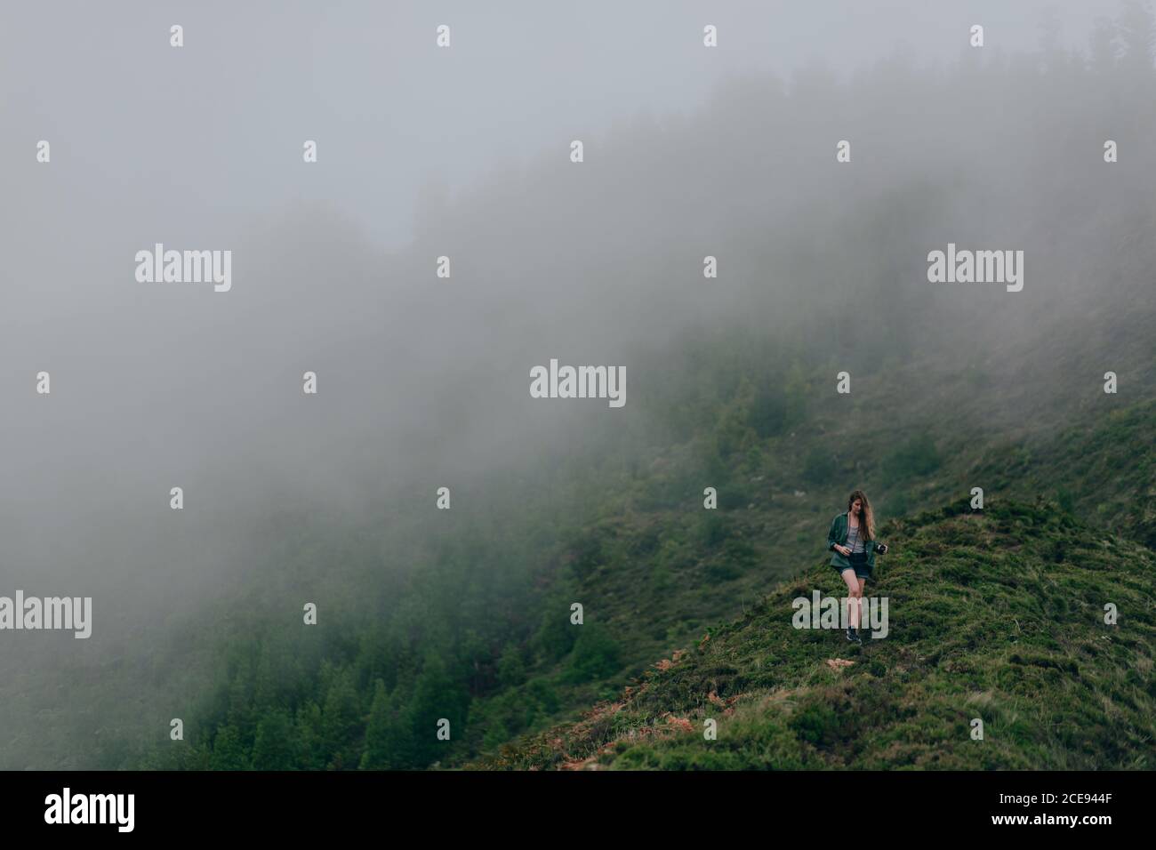 Donna che cammina su un'alta collina ripida coperta di erba verde con nebbia spessa sopra Foto Stock