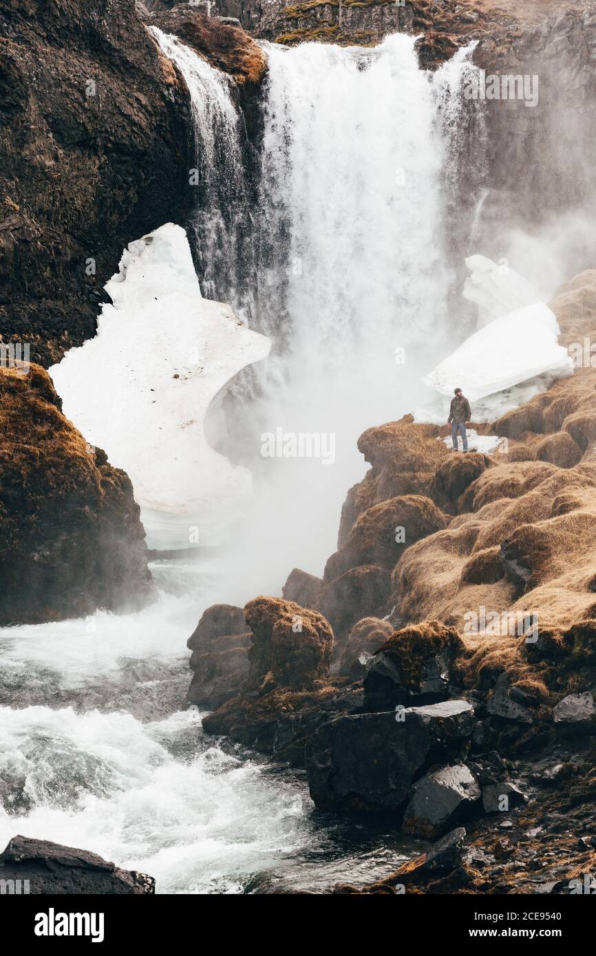 Vista a distanza di uomo in piedi sul bordo roccioso della collina con cascata che spruzzi sullo sfondo, Islanda. Foto Stock
