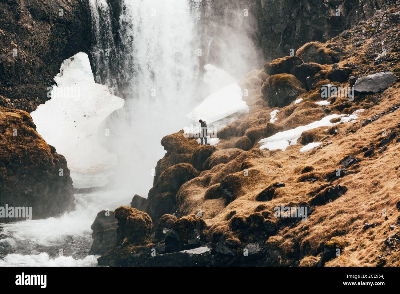Vista a distanza di uomo in piedi sul bordo roccioso della collina con cascata che spruzzi sullo sfondo, Islanda. Foto Stock