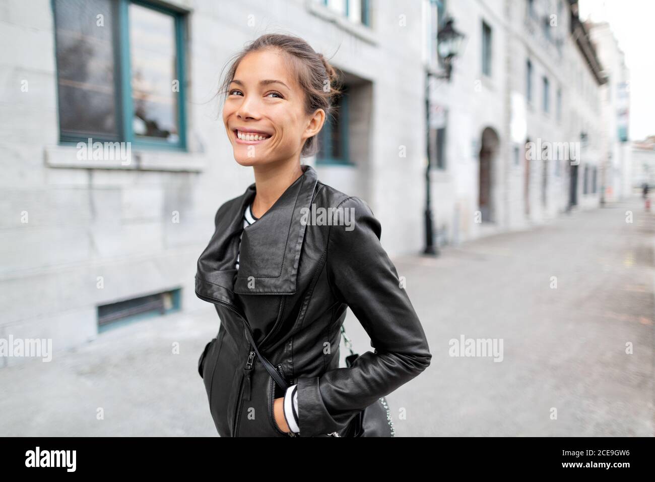 City Lifestyle People - giovane donna asiatica felice che cammina sulla vecchia strada del porto d'Europa sorridendo indossando giacca nera in pelle all'esterno. Urban elegante e alla moda Foto Stock