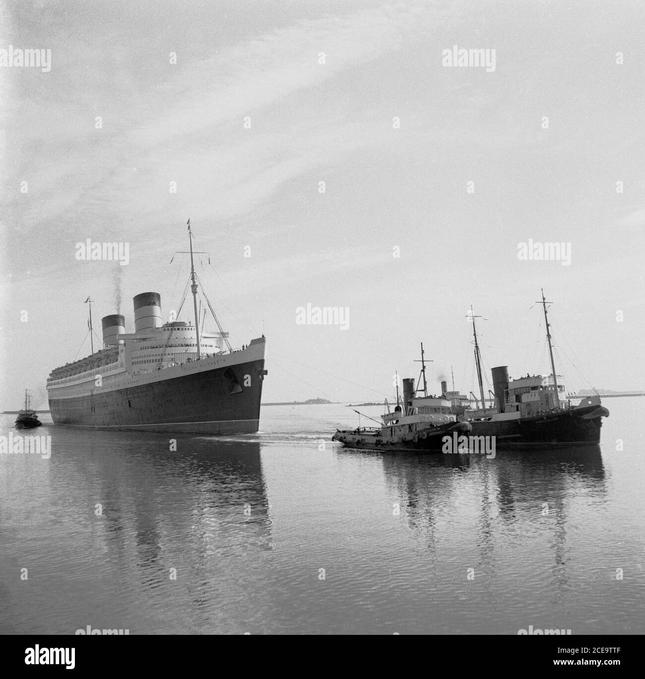 Anni '50, storici, tugboat tirare il RMS Queen Elizabeth transatlantico liner nelle acque calme del porto francese di Cherbourg. Prende il nome dalla regina madre britannica, era la più grande linea del passeggero mai costruita a quel tempo (1938) ed è rimasto così per 56 anni dopo. Fu usata come troopship in tempo di guerra durante la seconda guerra mondiale e divenne solo una nave di linea oceanica, come previsto, nel 1946. Da allora in poi, questo lussuoso liner ha fornito un servizio settimanale tra Southampton, Regno Unito e New York negli Stati Uniti, via Cherbourg, Francia. Nel 1972 a Victoria Harbour, Hong Kong, mentre subì una ristrutturazione, prese fuoco e affondò. Foto Stock