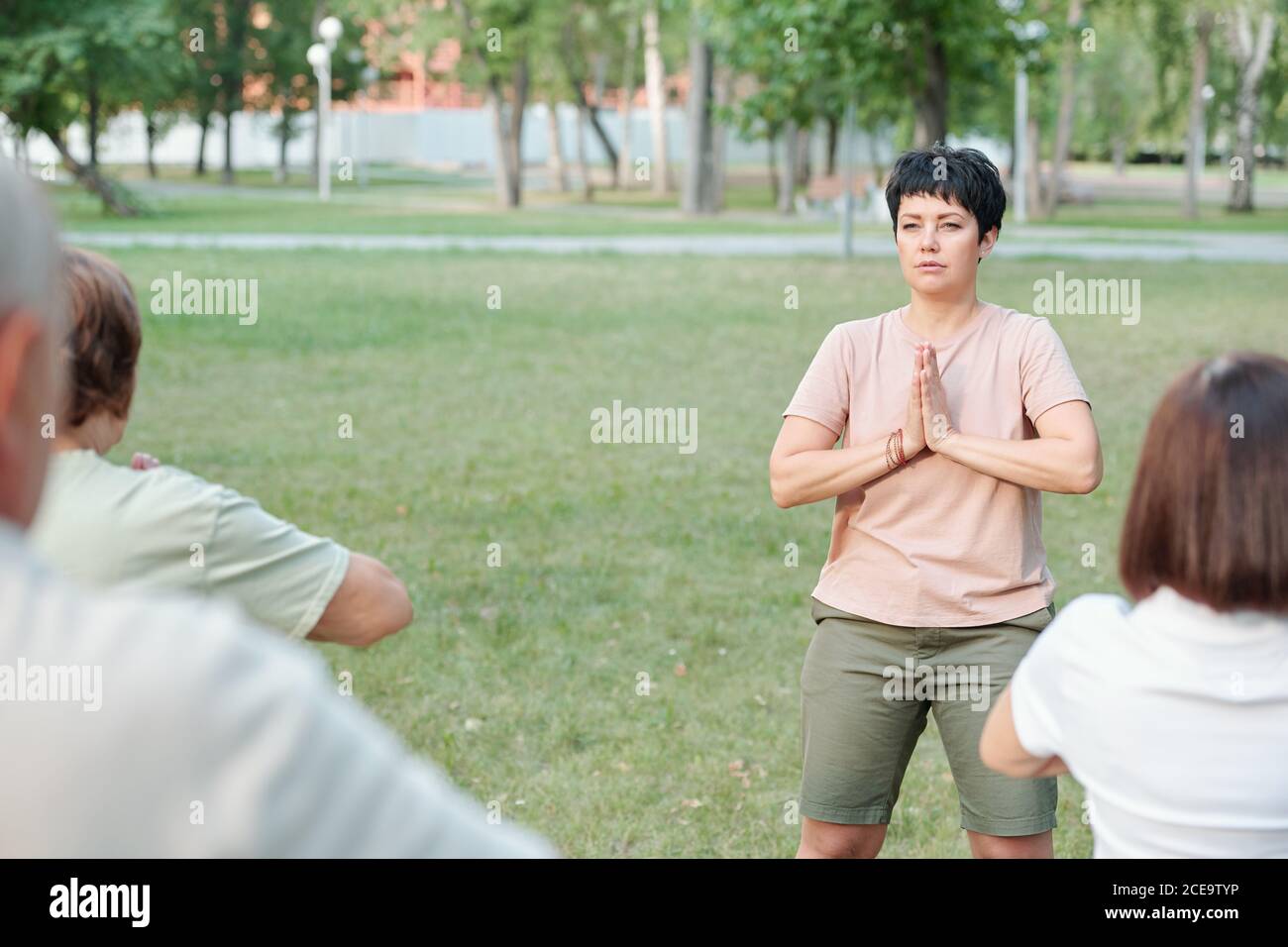 Insegnante di yoga focalizzata che fa gesto a Namaste mentre medita con gli studenti nel parco estivo Foto Stock