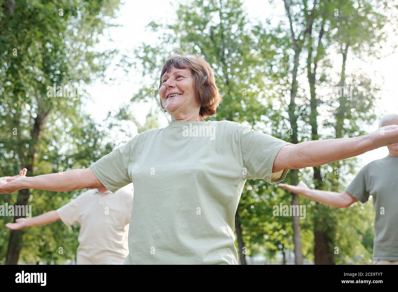Sotto la vista di allegra donna anziana energica in tshirt outstretching armi mentre si gode l'esercizio mattutino nel parco Foto Stock