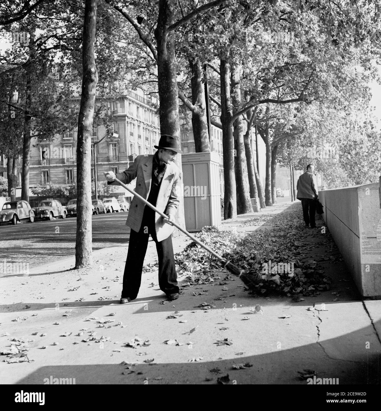Anni '50, storico, un uomo che indossa abiti civili e cappello spazzando su foglie caduti che giace su un marciapiede dagli alberi che fiancheggiano i viali vicino alla Senna a Parigi, Francia. Foto Stock