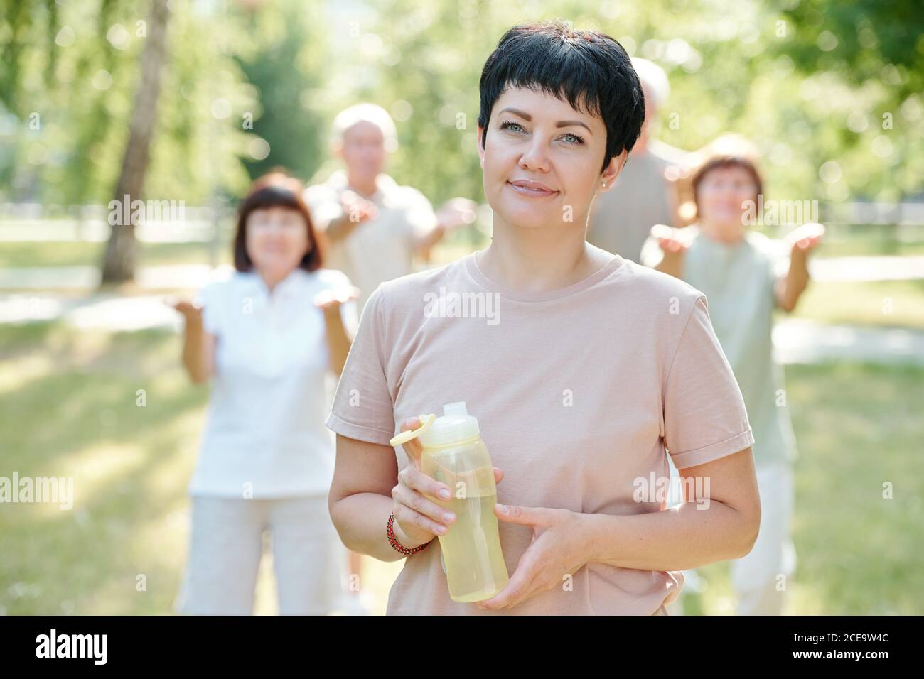 Ritratto di bruna sorridente allenatore fitness con capelli corti bottiglia d'acqua contro le persone anziane che fanno esercizio Foto Stock