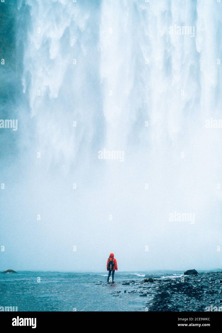 Vista posteriore di una persona irriconoscibile in giacca rossa con supporto per zaino contro la potente cascata. Islanda Foto Stock
