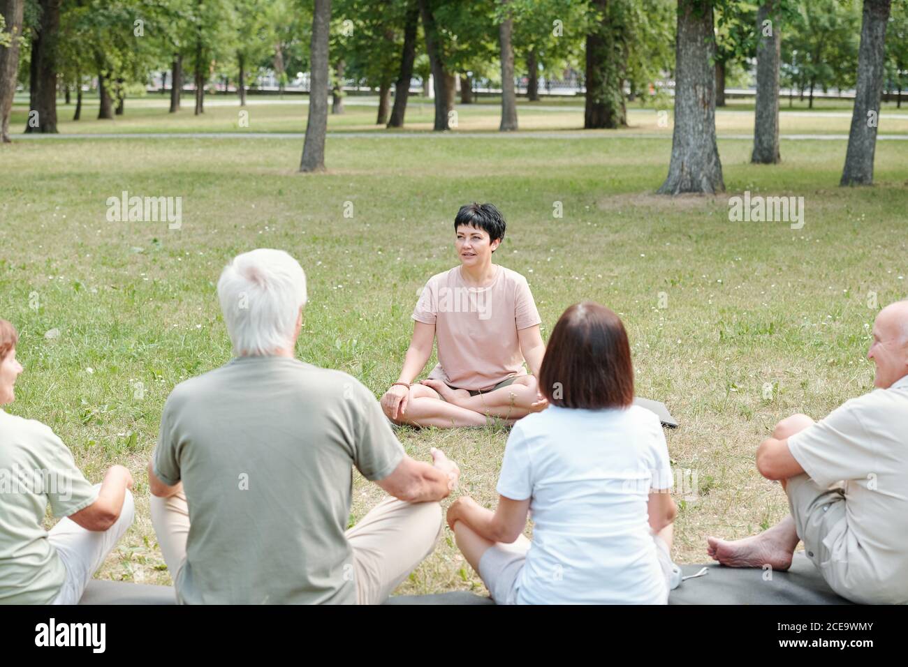 Allenatore professionale di yoga seduto in posizione lotus nel parco e. spiegare le basi dello yoga agli studenti più anziani Foto Stock
