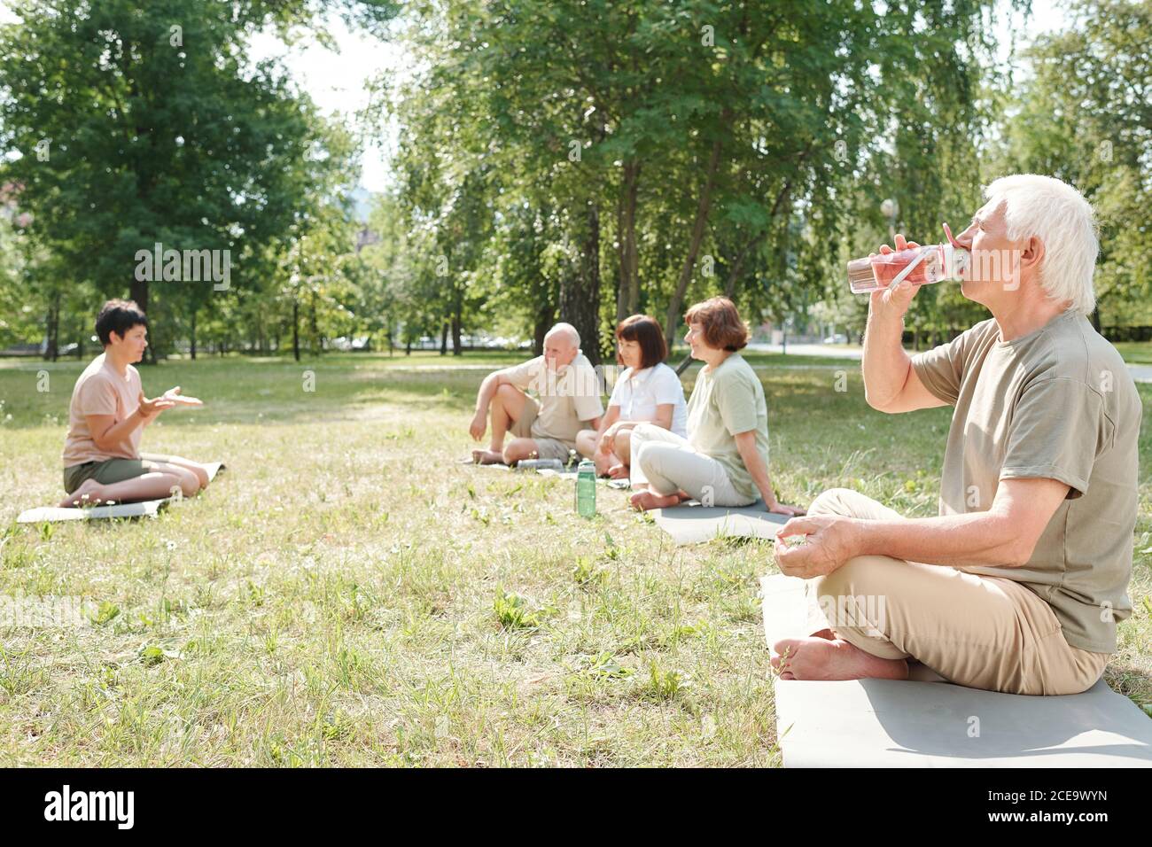 Uomo anziano con baffi seduti con gambe incrociate sul tappetino nel parco e nell'acqua potabile dopo la pratica di yoga Foto Stock