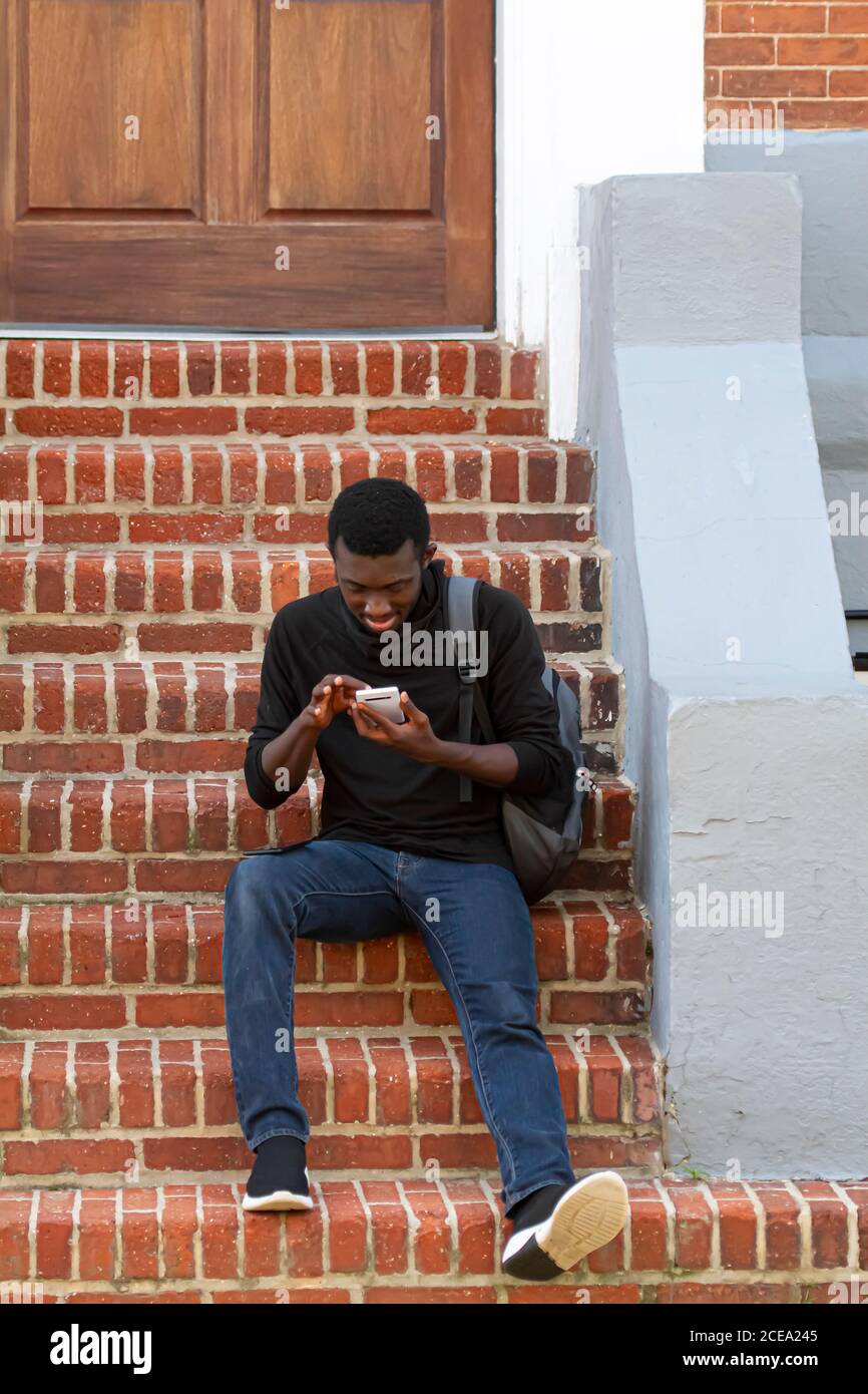 Annapolis, MD 08/21/2020: Un giovane afroamericano che indossa una t-shirt nera, jeans, sneakers e uno zaino è seduto su scale di mattoni fuori una h. Foto Stock