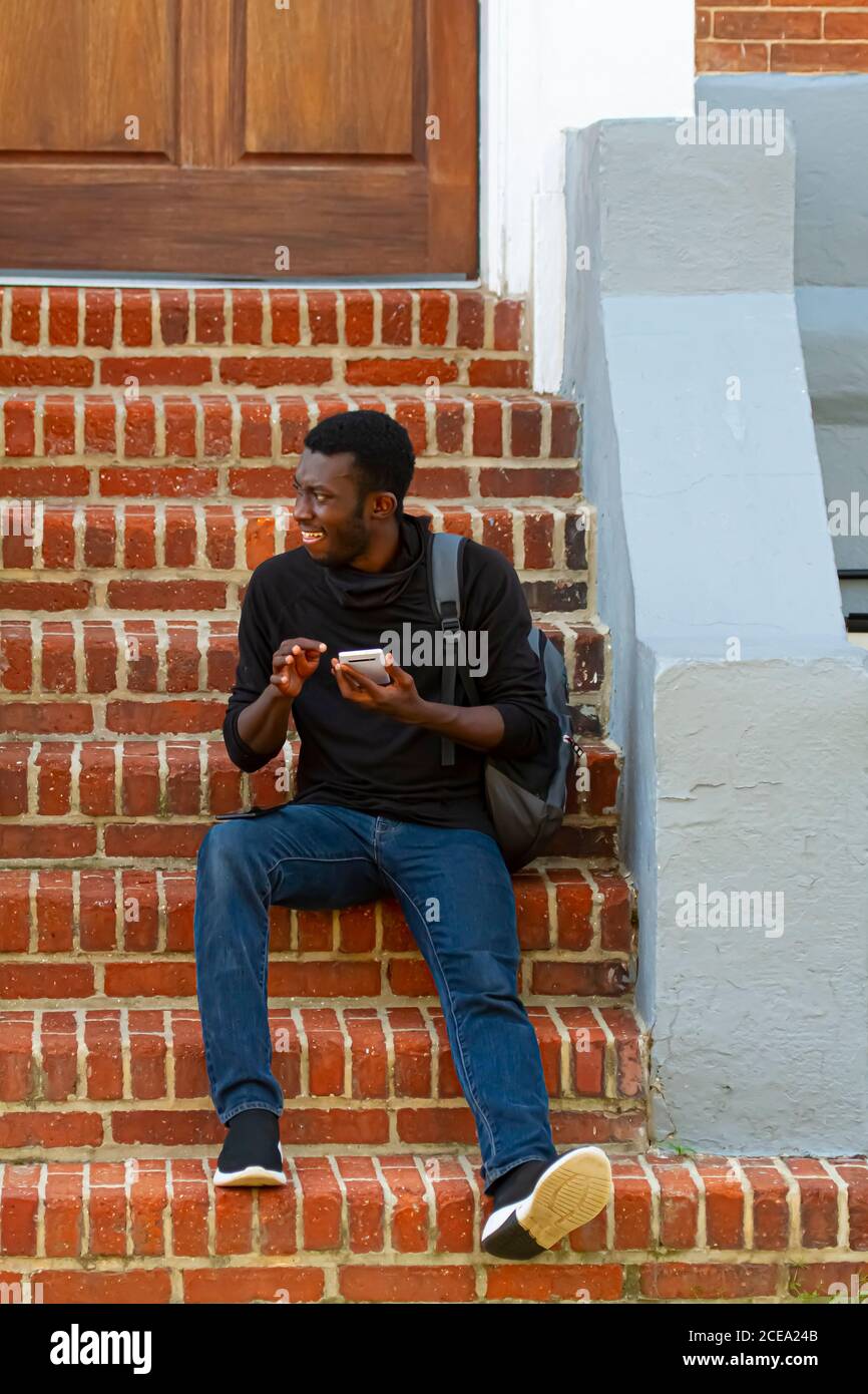 Annapolis, MD 08/21/2020: Un giovane afroamericano che indossa una t-shirt nera, jeans, sneakers e uno zaino è seduto su scale di mattoni fuori una h. Foto Stock
