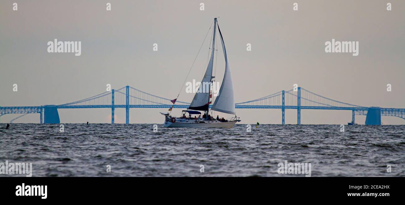 Immagine panoramica di una barca a vela che si muove attraverso la baia di Chesapeake con la sagoma del famoso Bay Bridge sullo sfondo. Ci sono persone sulla boa Foto Stock