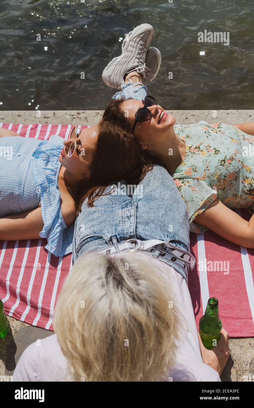 Dall'alto di belle donne in occhiali da sole godendo di sdraiarsi sopra gambe di un uomo alla moda che tiene in mano una bottiglia d'acqua Foto Stock