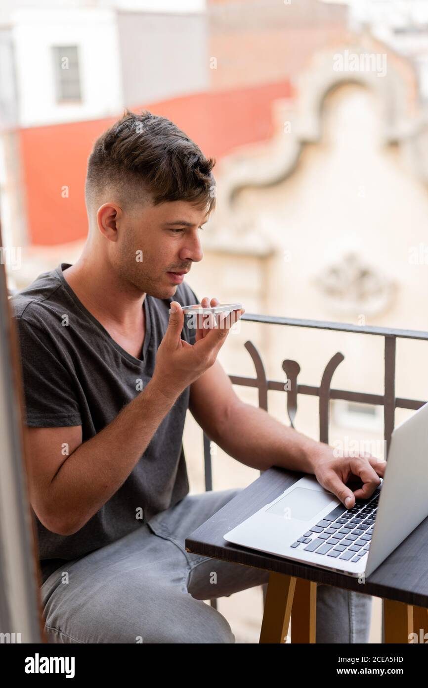 Adulto bel uomo seduto sul balcone guardando il computer portatile e l'invio messaggio vocale tramite smartphone Foto Stock