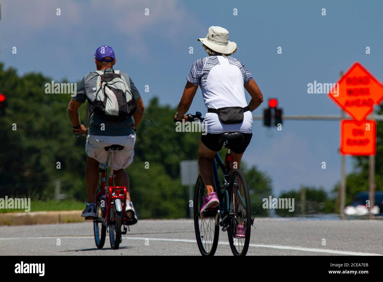 Una coppia anziana che indossa pantaloncini casual e t-shirt, così come i cappelli, è in bicicletta a cavallo vicino a un'autostrada in una soleggiata giornata estiva. L'uomo ha una pedalata pieghevole Foto Stock