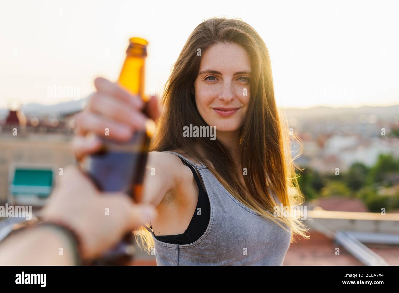 Vista del raccolto della mano che dà una bottiglia di birra fresca a. bella giovane donna in piedi sul tetto e guardando la macchina fotografica su sfondo retroilluminato sfocato Foto Stock