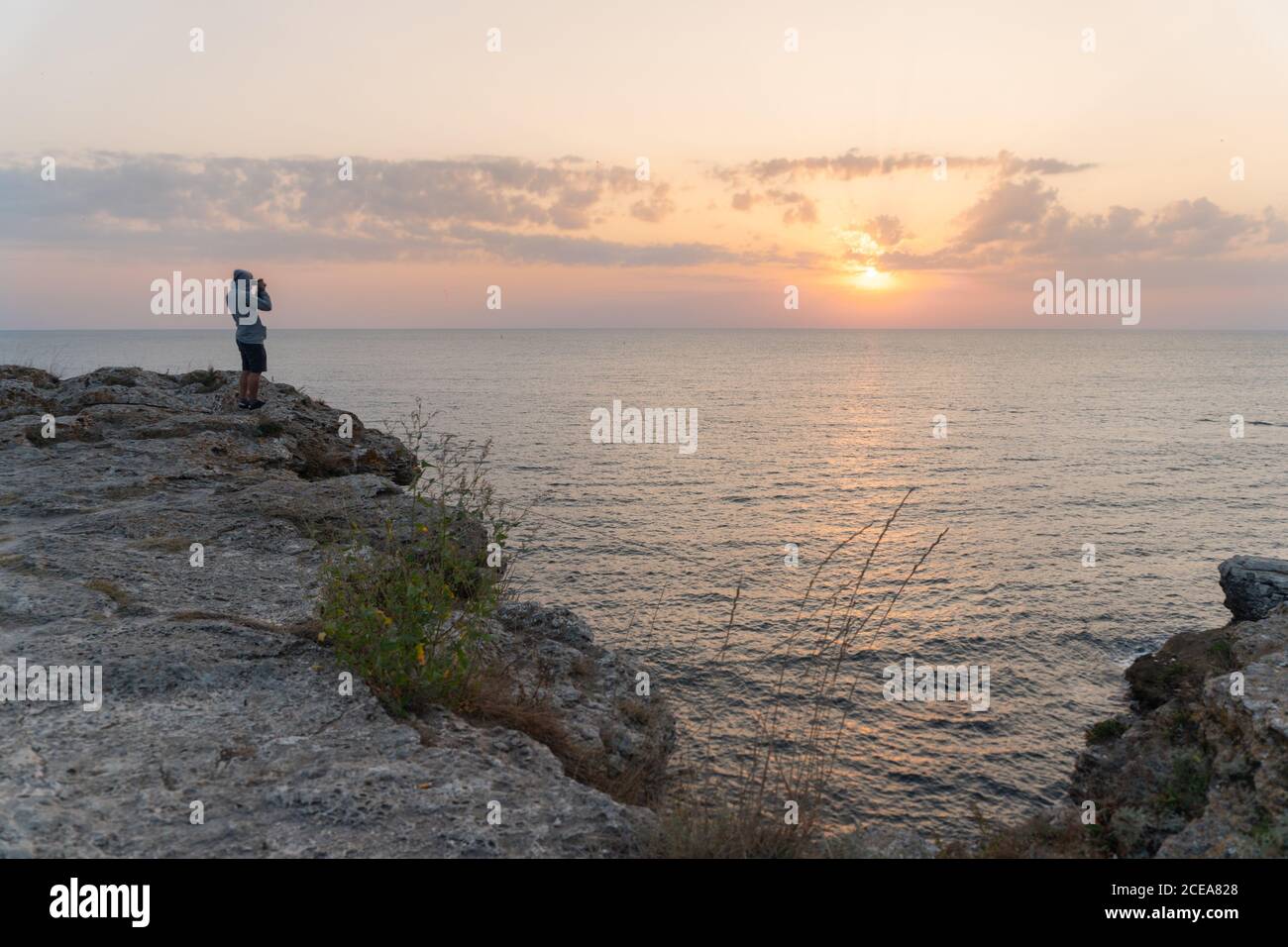 Vista laterale di un maschio irriconoscibile che si erge su una ruvida scogliera e che scatta foto del magnifico tramonto sul mare a Tyulenovo, Bulgaria Foto Stock
