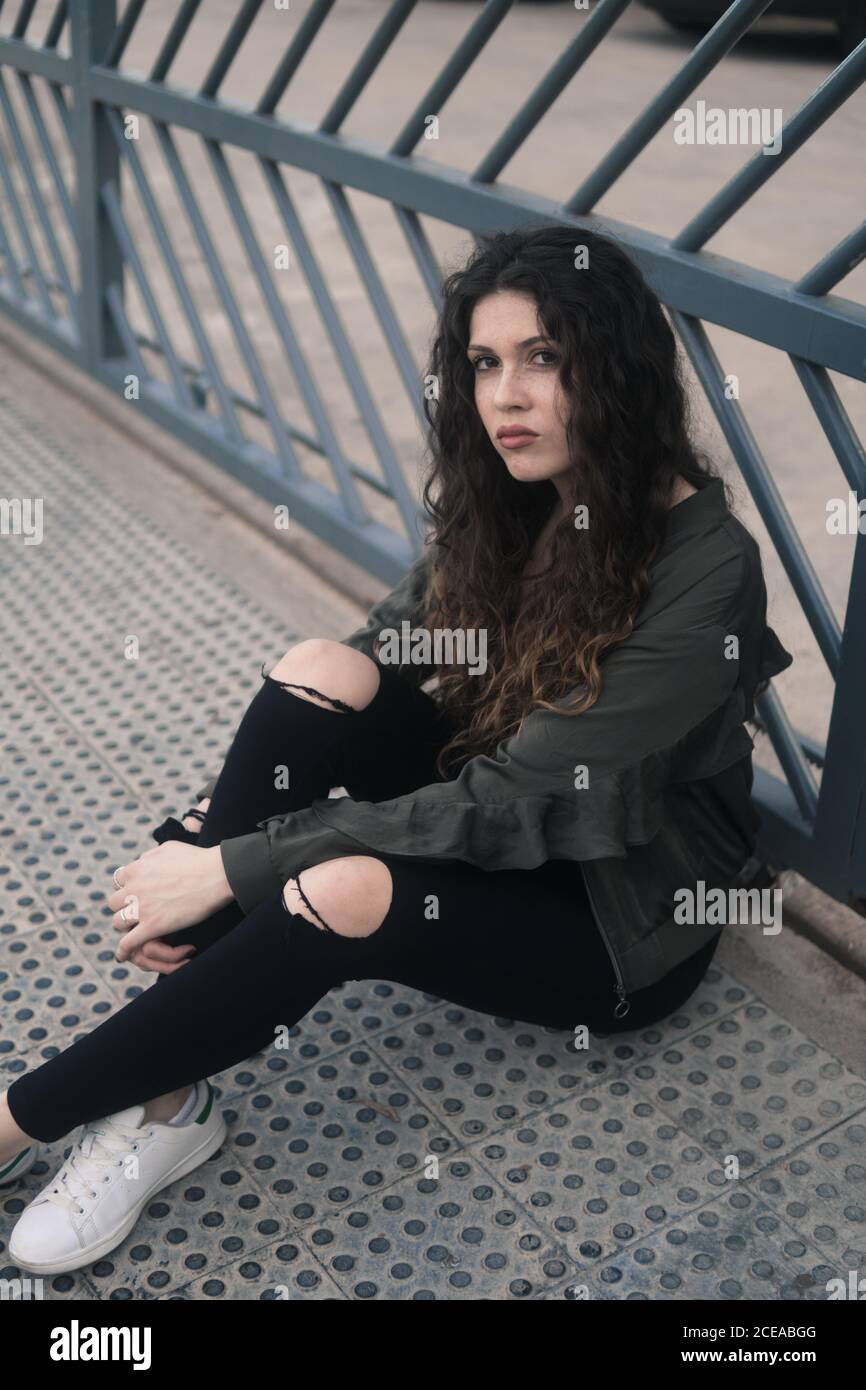 Bella giovane donna con capelli ricci guardando la macchina fotografica mentre si siede sul percorso metal alterato sulla strada cittadina Foto Stock