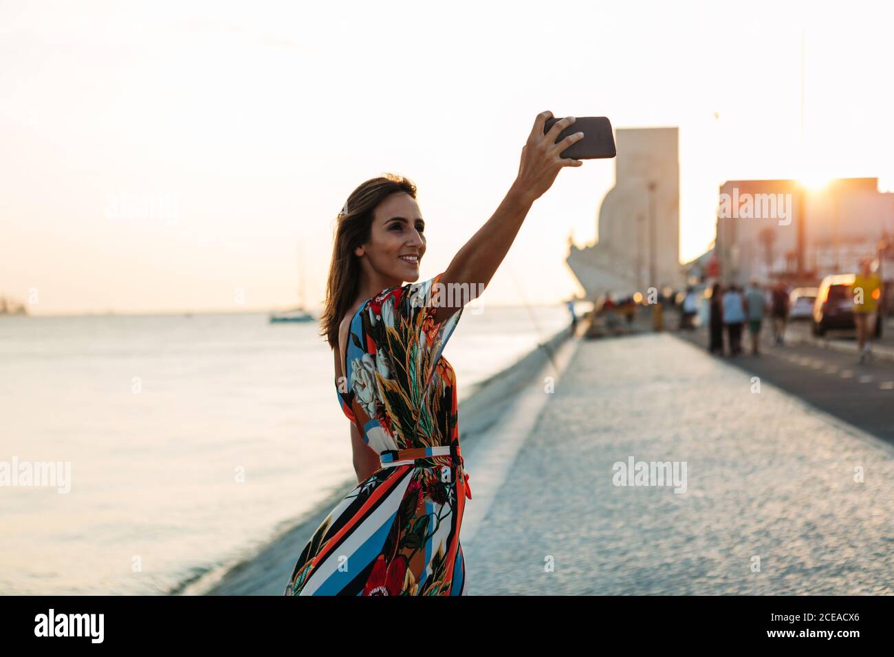 Donna soddisfatto nel vestito usando il telefono e prendendo selfie sopra lungomare al tramonto Foto Stock