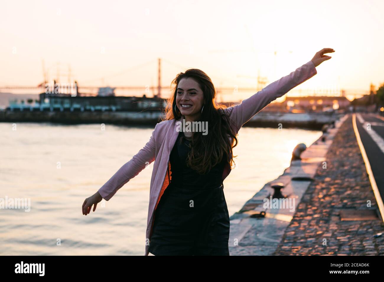 Sorridente passionale donna con la mano sui capelli in piedi argine vicino alla superficie dell'acqua al tramonto Foto Stock
