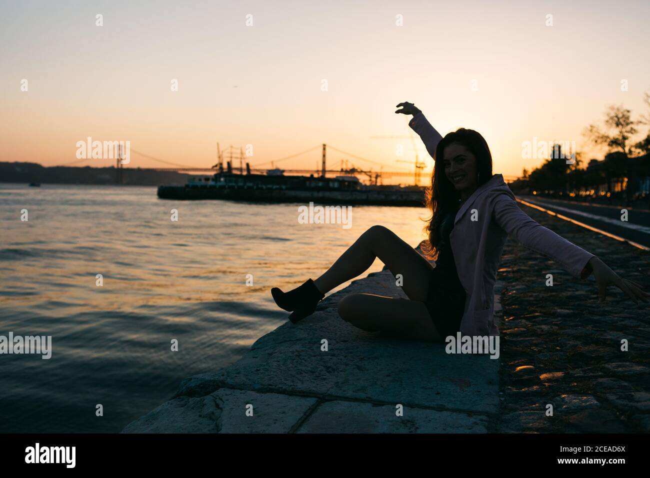 Vista laterale attraente signora passionale seduta sul terrapieno vicino all'acqua superficie con nave al tramonto Foto Stock