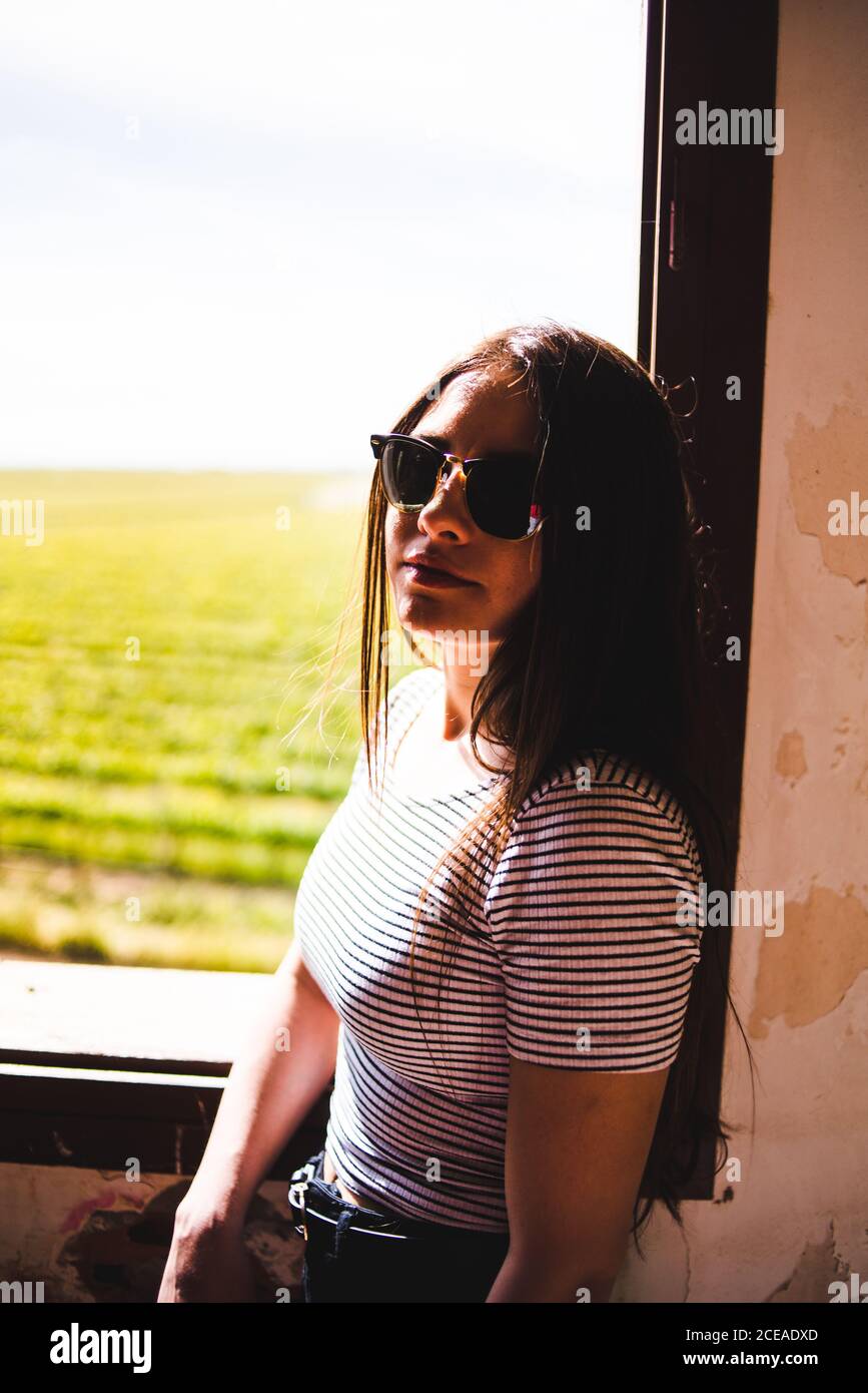 Giovane donna in occhiali da sole in piedi vicino alla finestra e guardando il bel campo verde nelle giornate di sole. Foto Stock