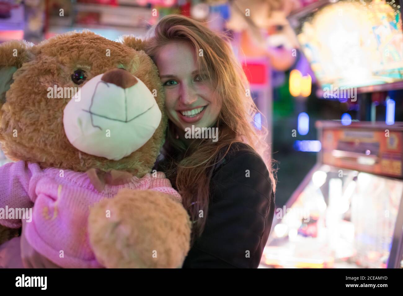 Donna allegra abbracciando orsacchiotto enorme guadagnato nel gioco macchina di divertimento e sorridendo alla macchina fotografica Foto Stock