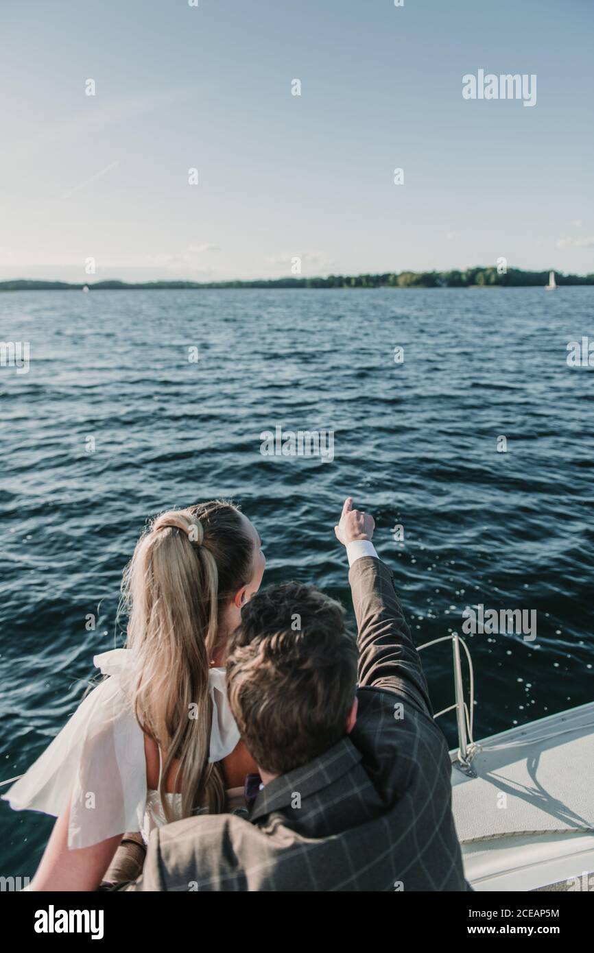 Vista posteriore dello sposo che punta via mentre si è in piedi con la sposa in barca sul lago Foto Stock