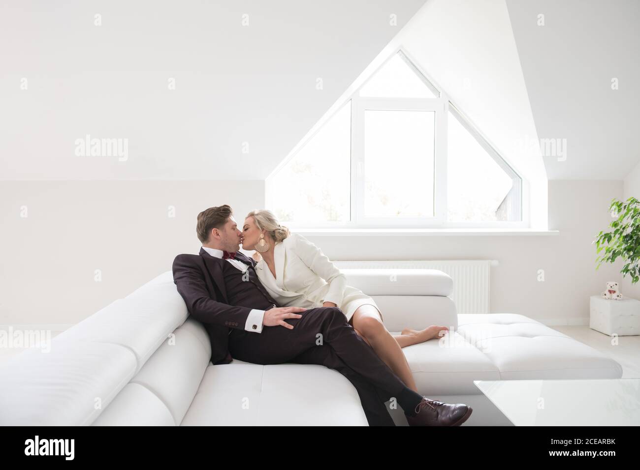 Vista di eleganti sposi moderni in un semplice bacio di interni bianchi sul divano in luce soffusa Foto Stock