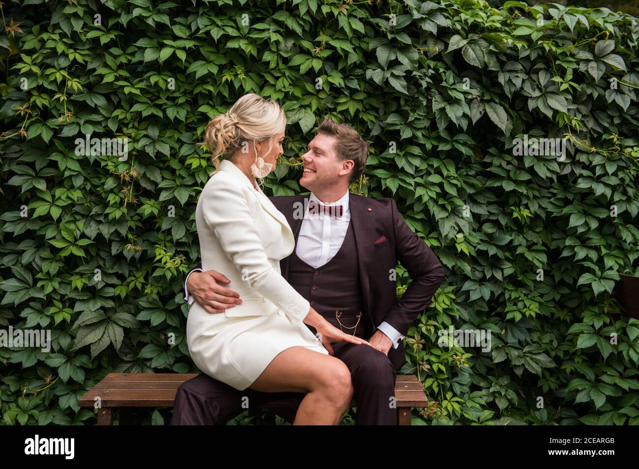 Uomo e donna eccitato indossando abiti da sposa alla moda e guardando l'un l'altro contro la siepe verde Foto Stock